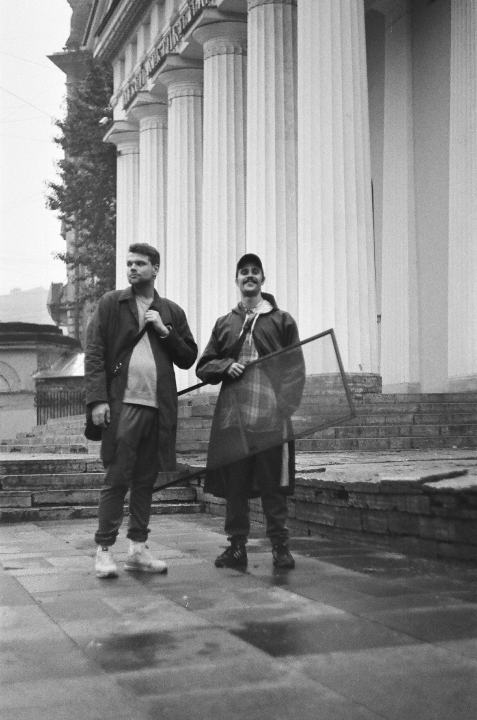 Black and white portrait of two men posing casually outdoors near a classical building.
